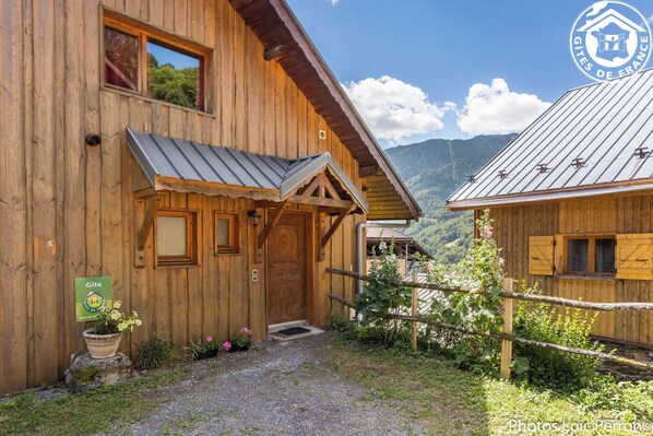 A wooden chalet with a metal roof, a wooden door, and a small porch. There is a sign on the door and a flower pot on the ground.