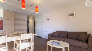 Una sala de estar moderna con un sofá marrón, una mesa de comedor con sillas y una mesa de centro. Hay luces colgantes rojas, un armario blanco y un cuadro en la pared.