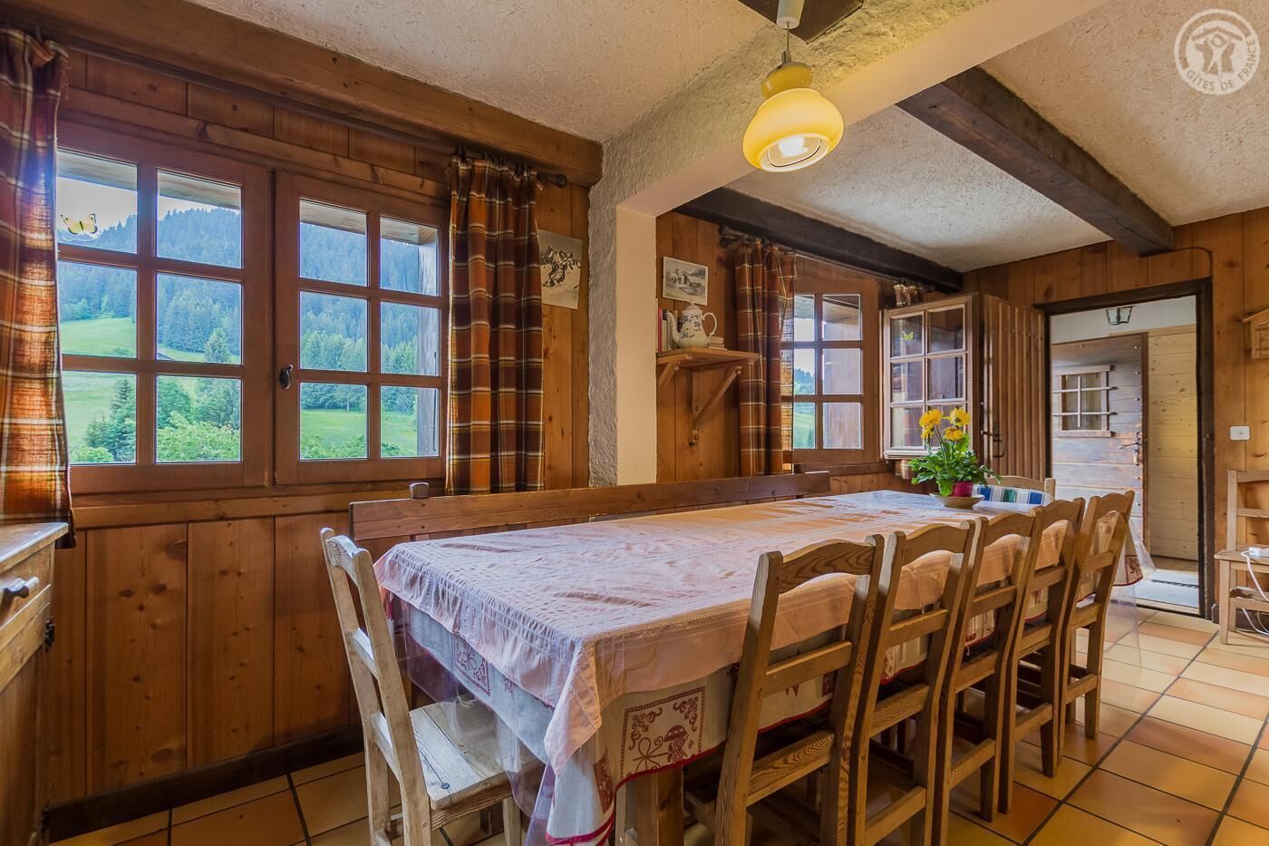 Ein Esszimmer mit Holzboden, einem Tisch für sechs Personen, flankiert von Fenstern mit Vorhängen, einer Hängeleuchte und Blick ins Grüne.