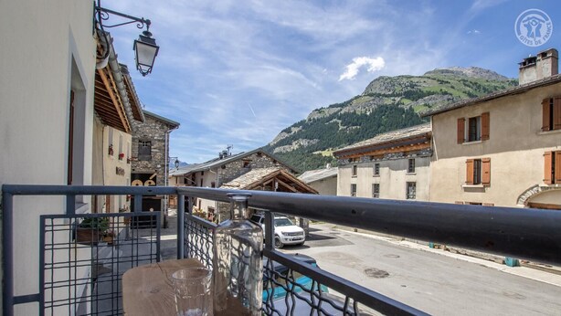 A balcony with a wooden table, a metal railing, and a view of a street with buildings and a mountain in the background. - LE BIVOUAC - AUSSOIS (AUSSOIS)