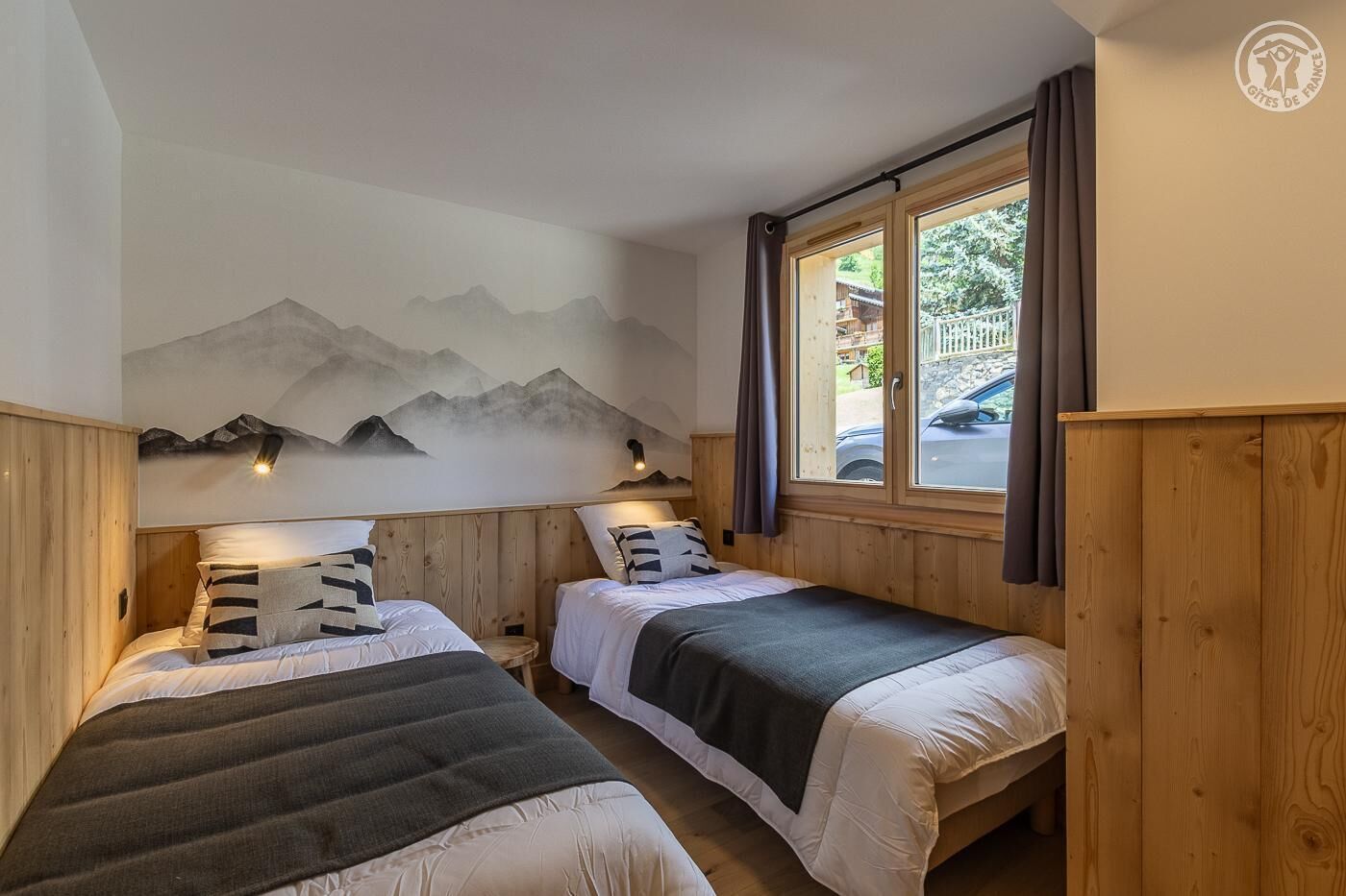 A room with two beds, wooden walls, a mountain mural, a window with curtains, and a door.