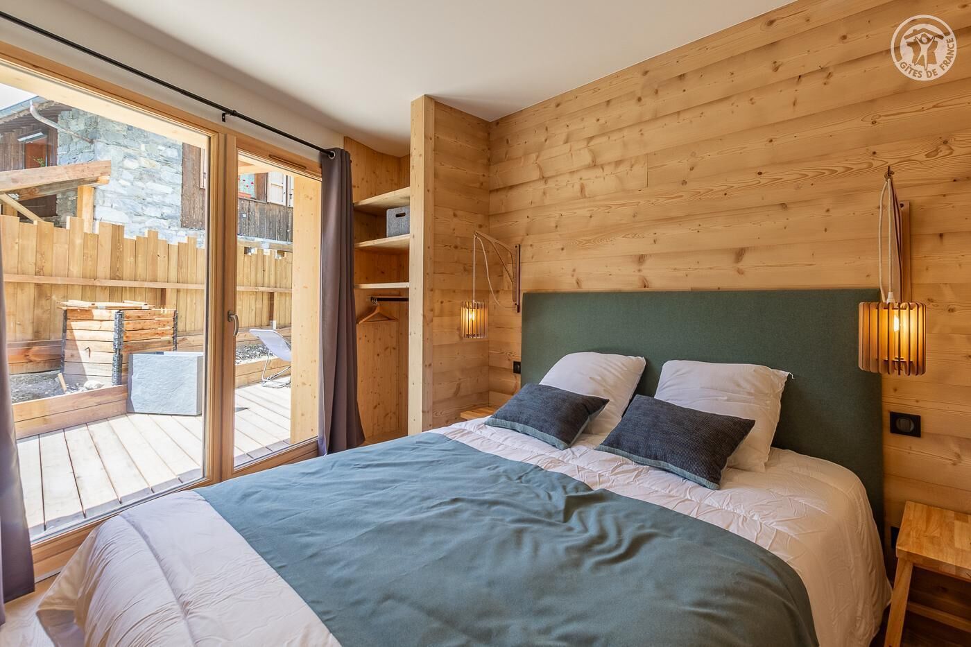 Ein Schlafzimmer einer Holzhütte mit einem Bett, zwei Nachttischen, einer Pendelleuchte und einer Schiebetür aus Glas, die zu einer Außenterrasse führt.