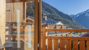 Una terraza de madera con un ventanal de vidrio que da a un cuarto con vista a una montaña y edificios tradicionales.