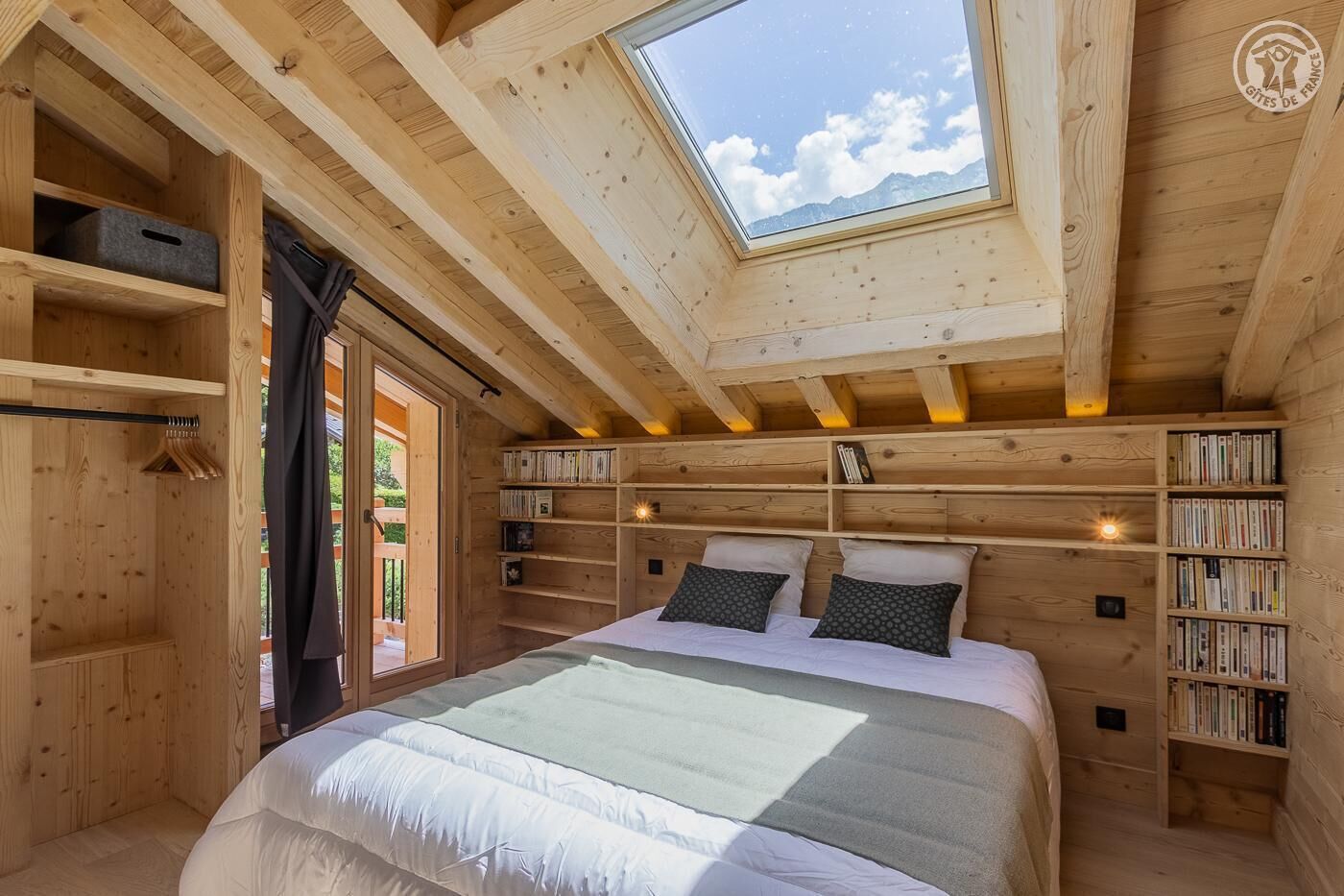A cozy wooden bedroom with a skylight, a bed with pillows, and built-in bookshelves.