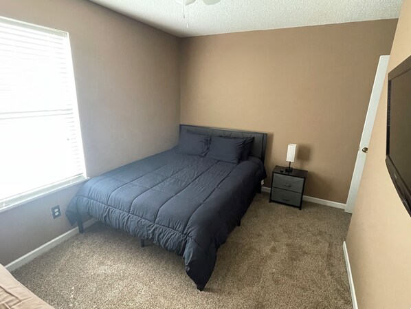 Une chambre avec un lit simple, une table de chevet, une lampe et une fenêtre avec des stores.