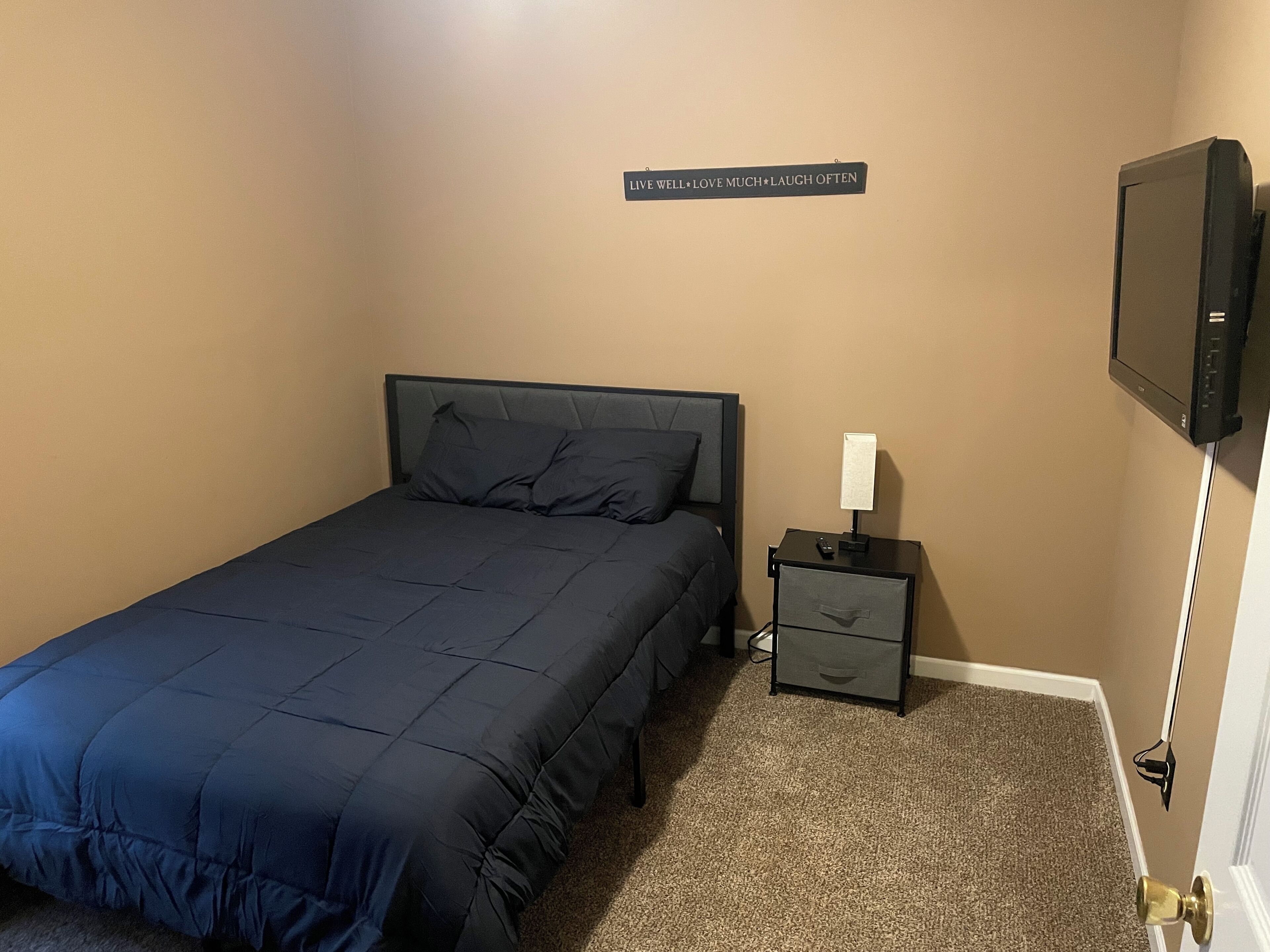 Un dormitorio con cama, mesita de noche, televisor y una frase motivadora en la pared.