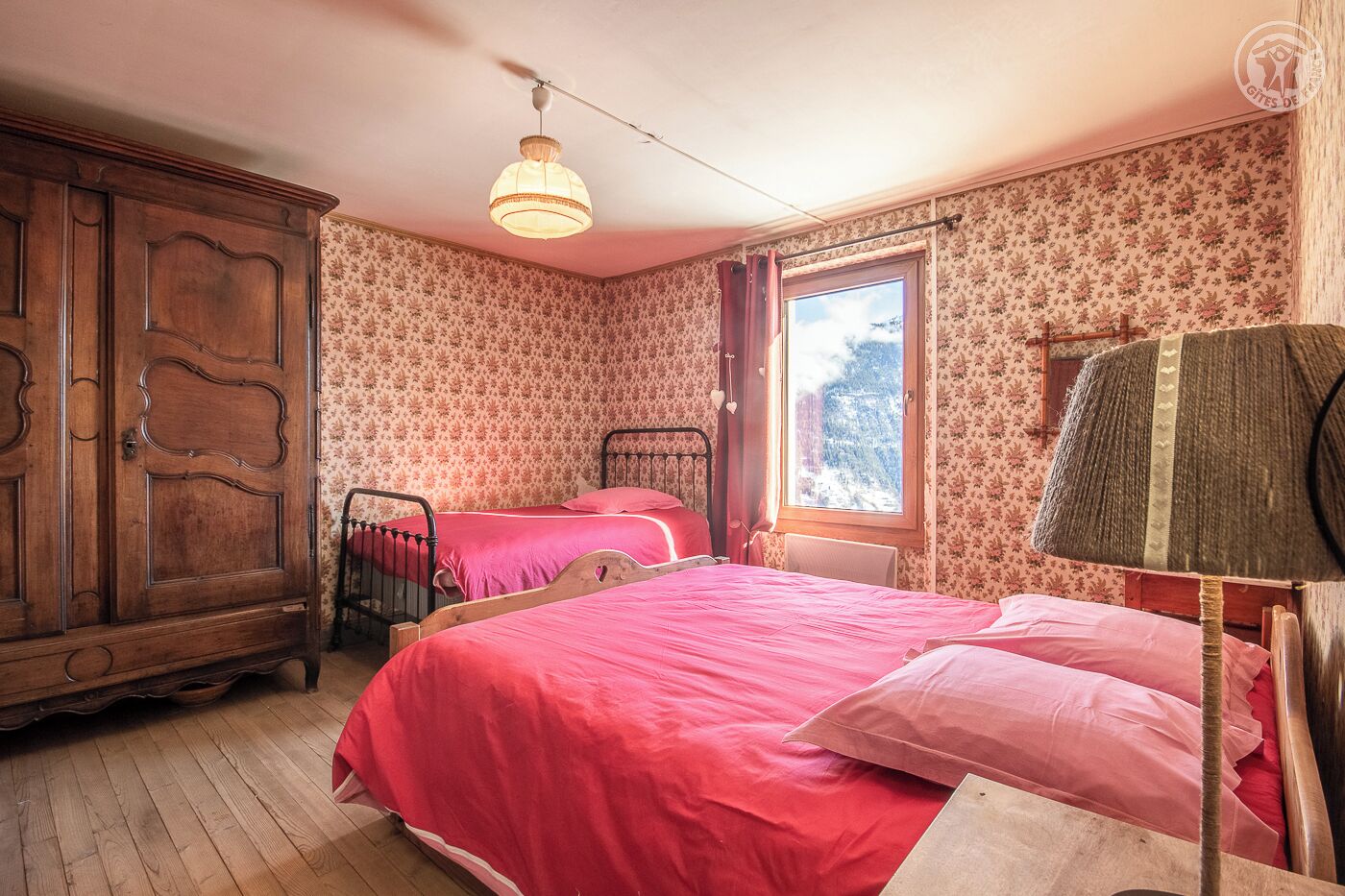 A bedroom with two beds, a wooden wardrobe, a window with a view of a snowy landscape, and a hanging lamp.
