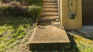 A house with a stone staircase leading to a wooden door, a garden with a neatly trimmed hedge, and a hose hanging on the wall. - LA PLANTAZ - CLERY - Gîte (CLERY)