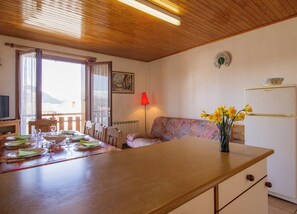 Uma sala de jantar com uma mesa de madeira posta para quatro pessoas, um sofá, um abajur vermelho e uma geladeira.
