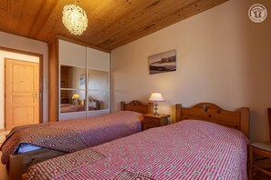 A room with two beds, wooden headboards, a wooden ceiling, a chandelier, a nightstand with a lamp, and a door. - LE CREY - CREST VOLAND COHENNOZ (CREST VOLAND COHENNOZ)