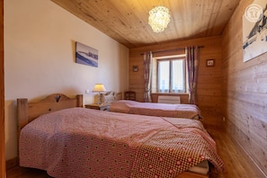 A room with two beds, a window with curtains, a wooden ceiling, and a picture on the wall.