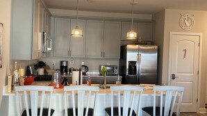 Een moderne keuken met lichtgrijze kastjes, roestvrijstalen apparatuur en een centraal eiland met witte stoelen.