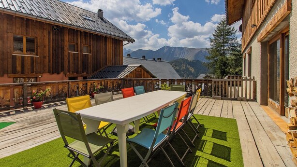 A wooden deck with a table and chairs, surrounded by mountains and a clear sky.