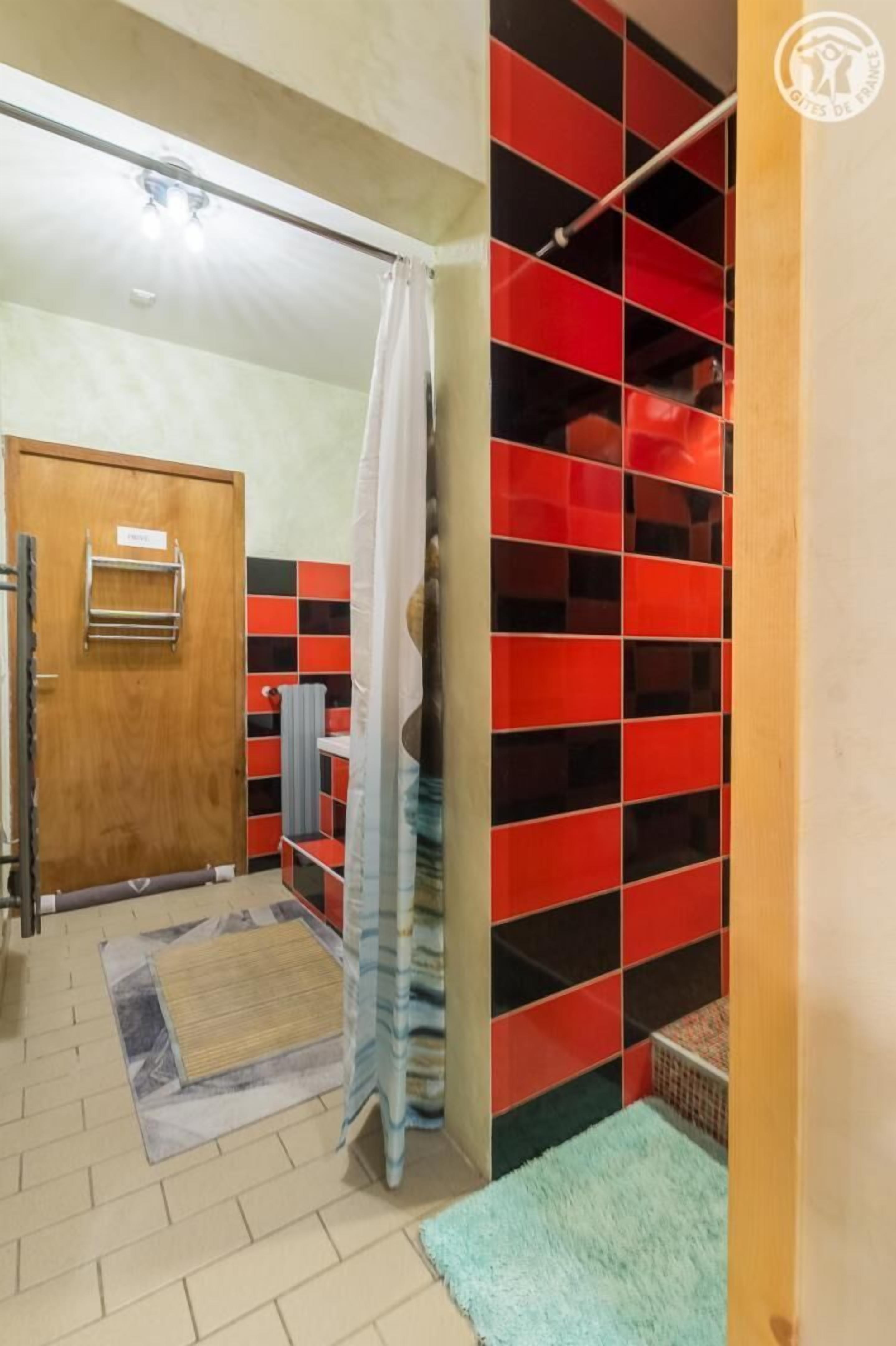 A bathroom with a wooden door, a towel rack, a shower curtain, and a tiled wall with red and black tiles.