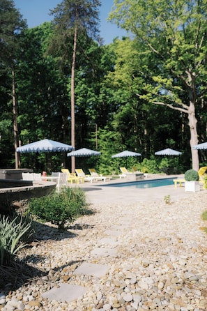 Un espace piscine agrémenté de chaises longues et de parasols, entouré d’arbres.