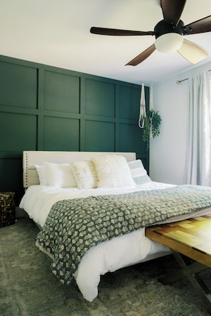 A bedroom with a bed, a ceiling fan, a hanging plant, and a window with curtains.