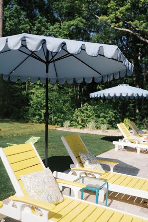 Buitenloungestoelen met parasols, geplaatst op een terras met gazon en bomen op de achtergrond.