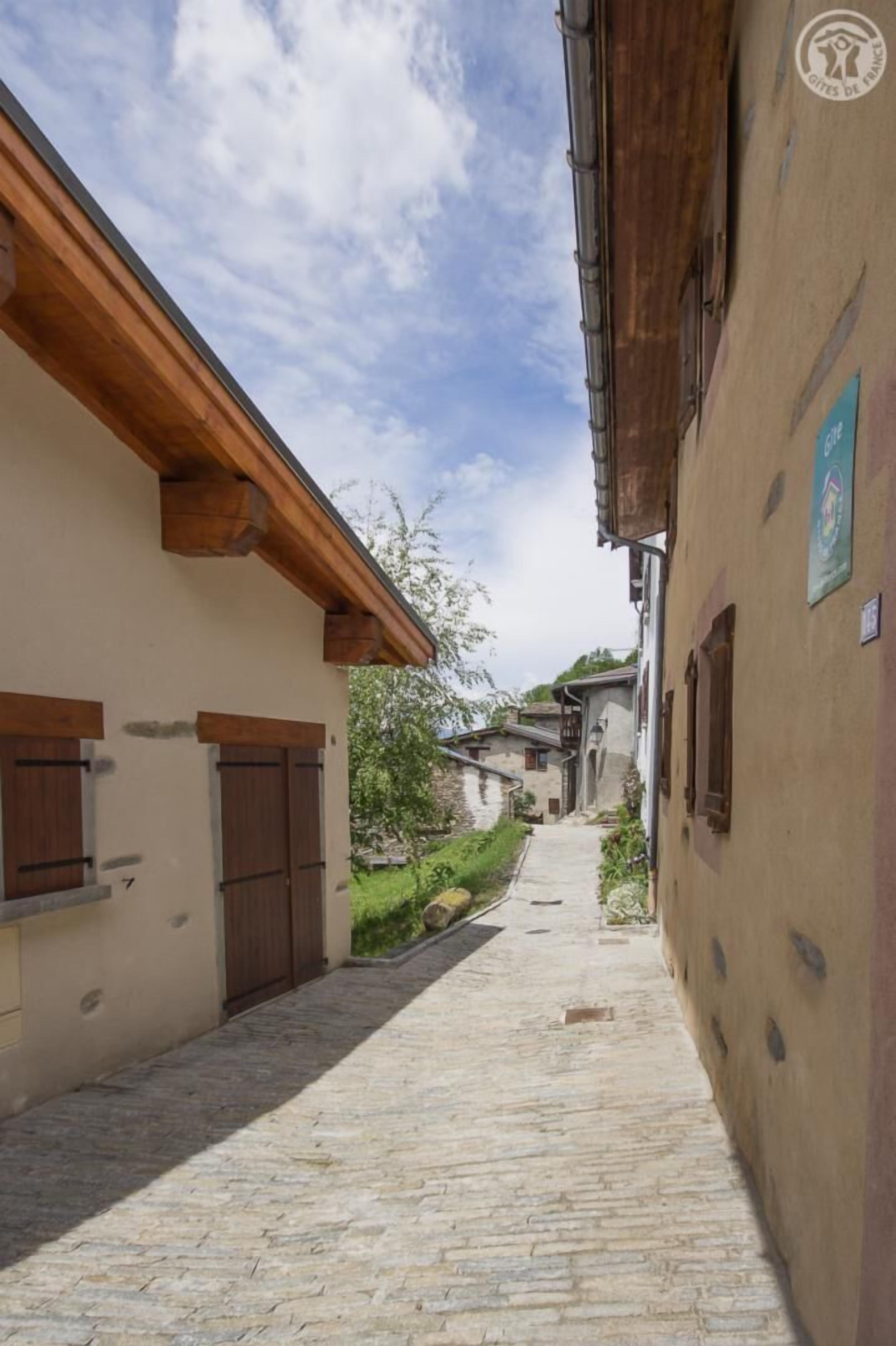 En smal brostensgade omgivet af traditionelle huse med træpersienner og en klar, blå himmel.