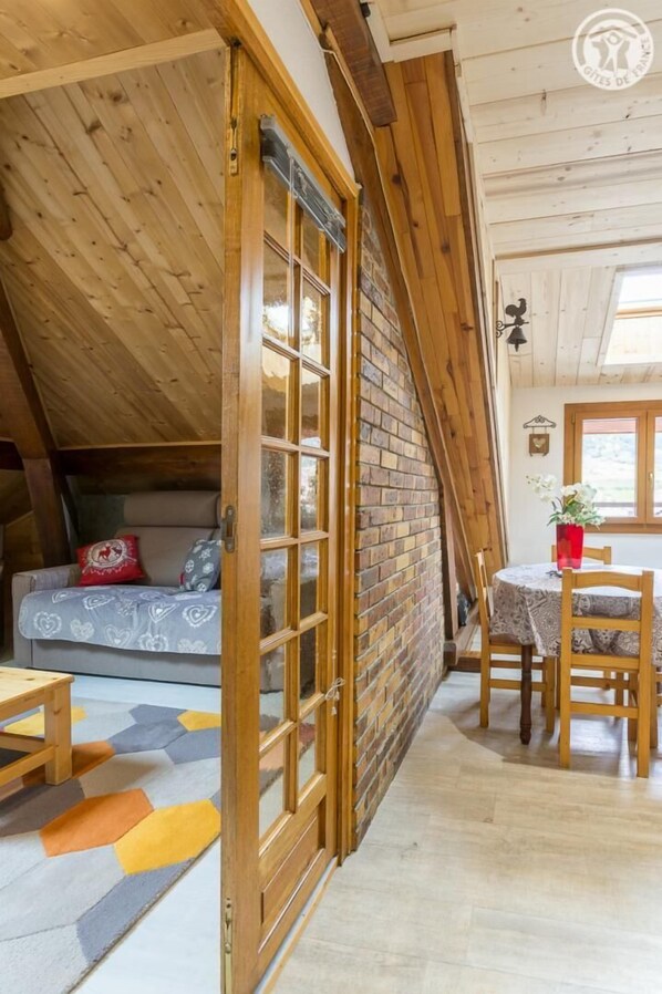 A cozy attic bedroom with a wooden sloped ceiling, a brick wall, a wooden door, a sofa with cushions, a dining table with chairs, and a window with a view.