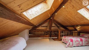Ein Schlafzimmer im Dachgeschoss mit hohen Decken, Holzbalken, einem Oberlicht und einem Bett mit gemustertem Bettbezug.