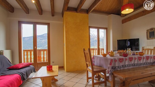 Ein gemütliches Wohnzimmer mit einer Couch, einem Holztisch und einem Esstisch für zwei Personen. Große Fenster bieten Blick auf die Berge, es gibt einen Fernseher und eine rote Lampe.
