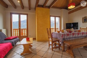 Ein gemütliches Wohnzimmer mit einer Couch, einem Holztisch und einem Esstisch für zwei Personen. Große Fenster bieten Blick auf die Berge, es gibt einen Fernseher und eine rote Lampe.