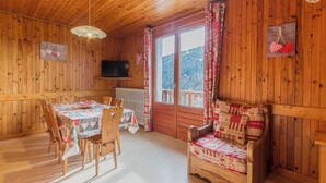A wooden cabin with a dining table and chairs, a sofa, a television, and a view of the forest through the windows.