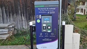An electric vehicle charging station with a sign indicating it is for 'SAUF' vehicles, surrounded by a rustic wooden fence and a stone wall.
