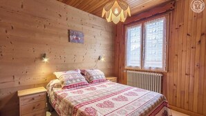 A wooden cabin bedroom with a bed, bedside tables, a hanging light fixture, and a window with curtains.