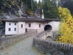 Uma ponte de pedra que leva a um edifício branco, com um muro de pedra de um lado e uma área arborizada do outro.