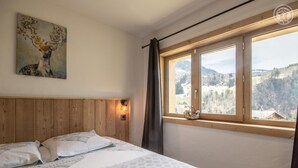 Une chambre à coucher avec une tête de lit en bois, un lit recouvert d’une literie à motifs, un luminaire fixé au mur et une fenêtre donnant sur un paysage montagneux.