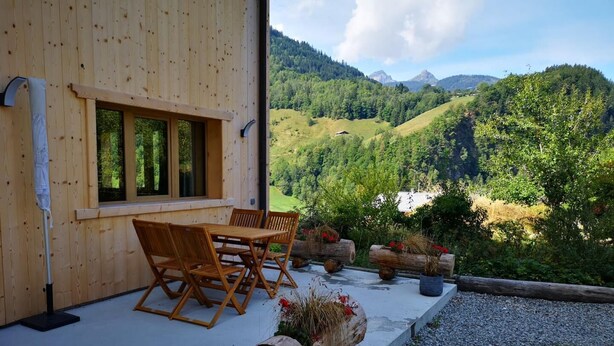 Une cabane en bois dotée d’une terrasse avec une table et des chaises, de jardinières en rondins de bois remplies de fleurs et offrant une vue sur un paysage montagneux.