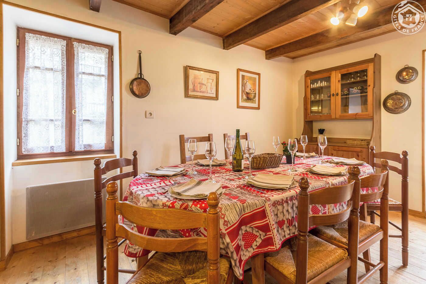 A dining room with a wooden table set for four, surrounded by wooden chairs. There is a window with lace curtains, a wall-mounted pan, and framed pictures above the table.