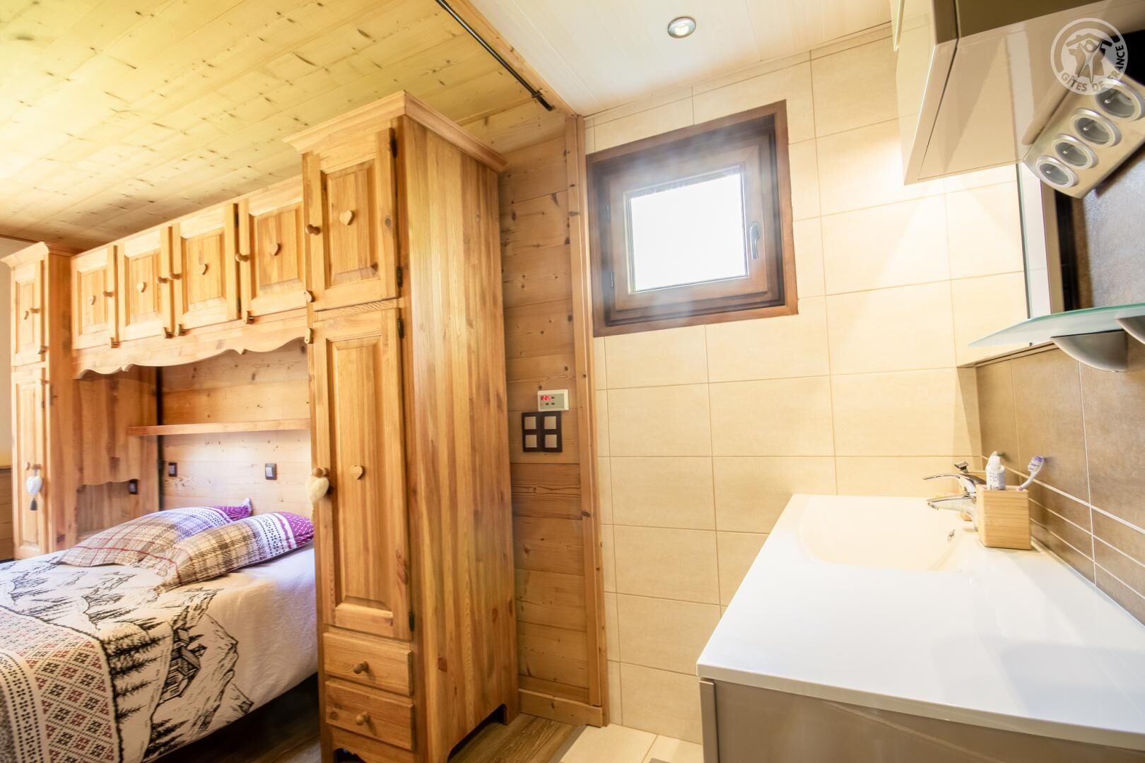 Ein holzgetäfeltes Zimmer im Blockhausstil mit einem Bett, einem Holzschrank, einem kleinen Fenster und einem Badezimmer mit Waschbecken und Spiegel.
