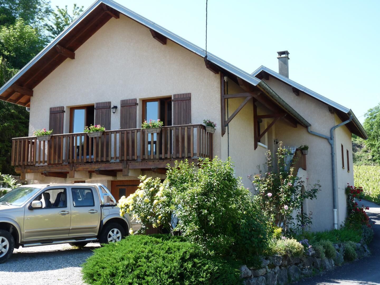 A two-story chalet with a balcony, a car parked in front, and lush greenery around.