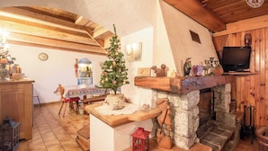 A cozy, rustic interior with a stone fireplace, wooden beams, a small Christmas tree, and a television mounted on the wall.