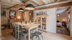 A rustic dining area with a wooden table set for four, surrounded by metal chairs. A wooden cabinet with glass doors is visible in the background, and a bed is seen in the adjacent room.