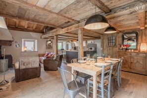 Un interior rústico con vigas de madera a la vista, una zona de comedor con mesa de madera y sillas de metal, una cocina con electrodomésticos modernos y un rincón de estar acogedor con sofá y cojines.