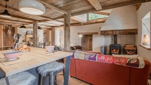 A rustic interior with a wooden ceiling, a fireplace, a dining area with a table and chairs, and a sofa with a colorful blanket.