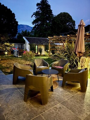 Um pátio externo com cadeiras e uma mesa, rodeado por plantas e uma pérgula com luzes de cordão.