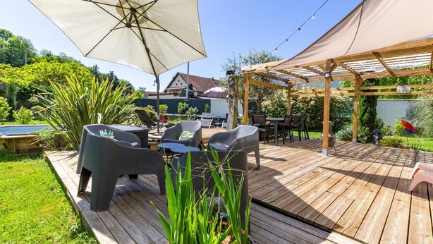 Un terrazzo in legno con arredi da esterno, un pergolato e una piscina sullo sfondo.