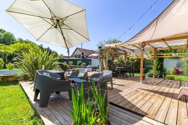A wooden deck with outdoor furniture, a pergola, and a swimming pool in the background.