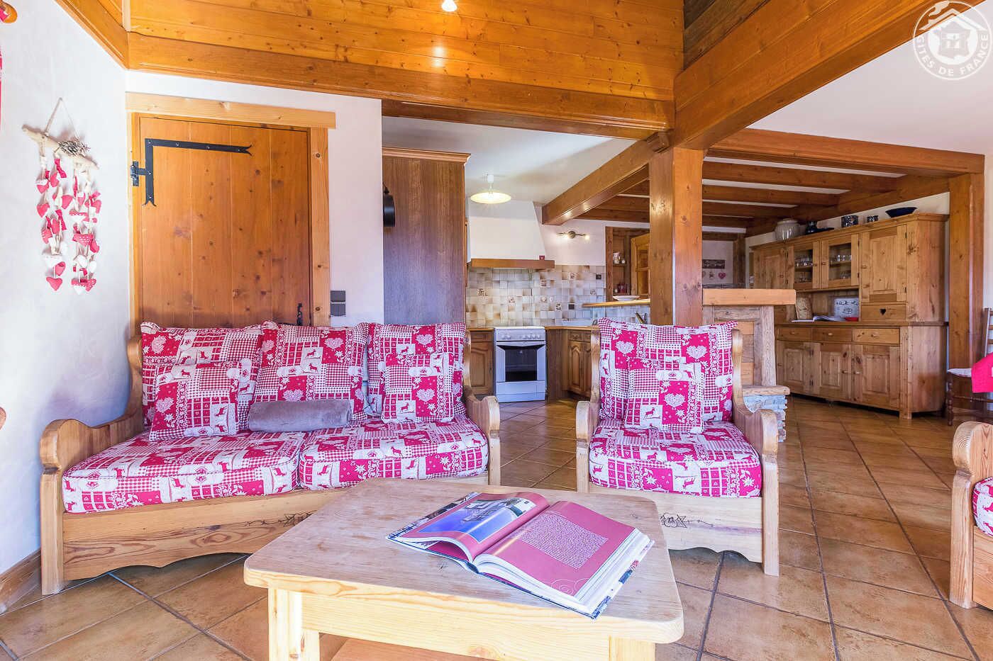 Un salon chaleureux avec un canapé et des fauteuils à motifs, une table basse en bois sur laquelle repose un livre, et une cuisine en arrière-plan.