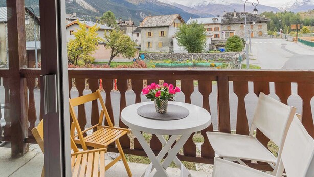 A balcony with a table and chairs, overlooking a mountain village with traditional buildings.