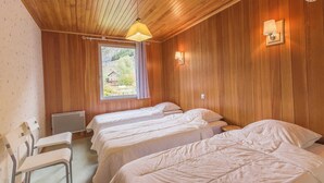 A room with wooden walls and ceiling, two beds with white bedding, a window with a view of greenery, and a wall-mounted lamp.
