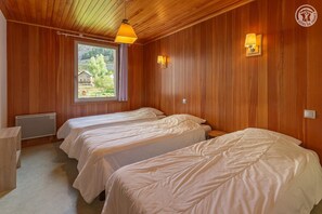A room with wooden walls, two beds with white bedding, a window with a view of greenery, and a hanging light fixture.