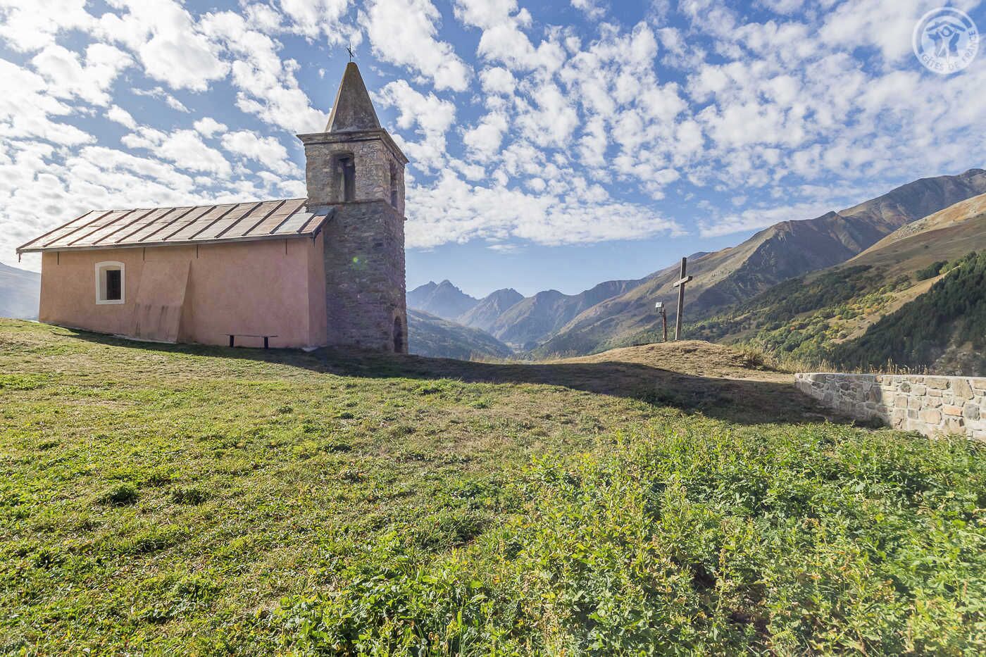 A small stone church with a bell tower and a cross on a hill, surrounded by mountains and greenery.