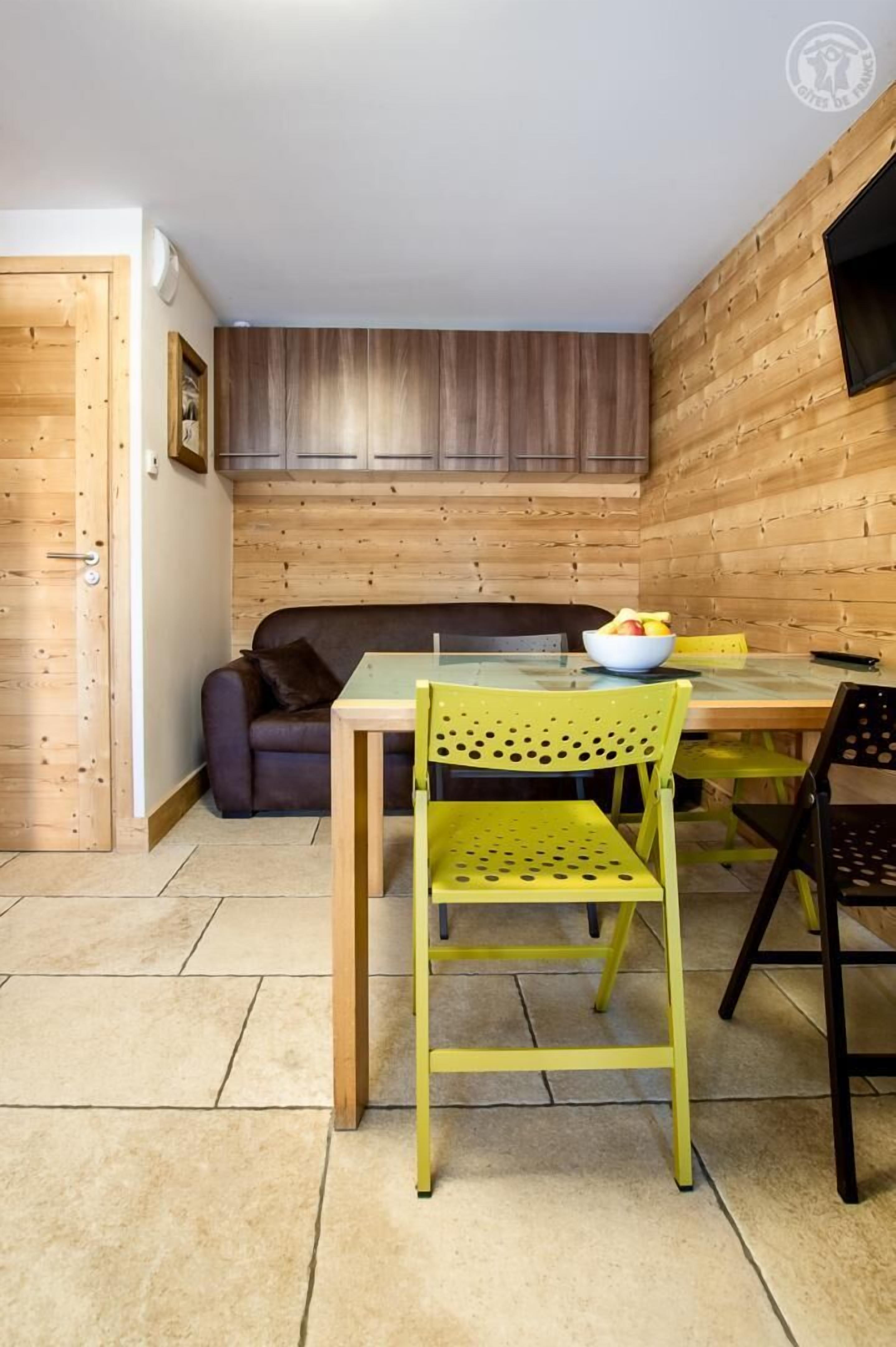 A compact living space with a wooden wall, a sofa, a dining table with chairs, and a bowl of fruit on the table.