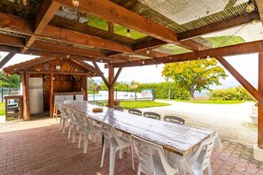 A covered outdoor dining area with a long wooden table and white plastic chairs, a refrigerator, and a view of a swimming pool and greenery.