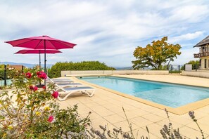 A swimming pool with lounge chairs and a red umbrella, surrounded by greenery and a view of the landscape.